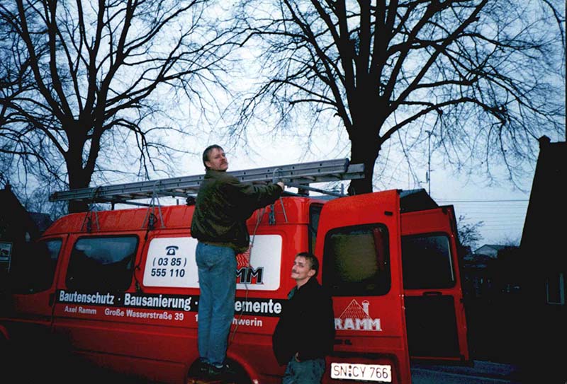 Altes Foto aus den Anfängen: Axel Ramm steht links auf dem roten Firmenwagen und macht eine Leiter fest, rechts daneben ein Kollege.
